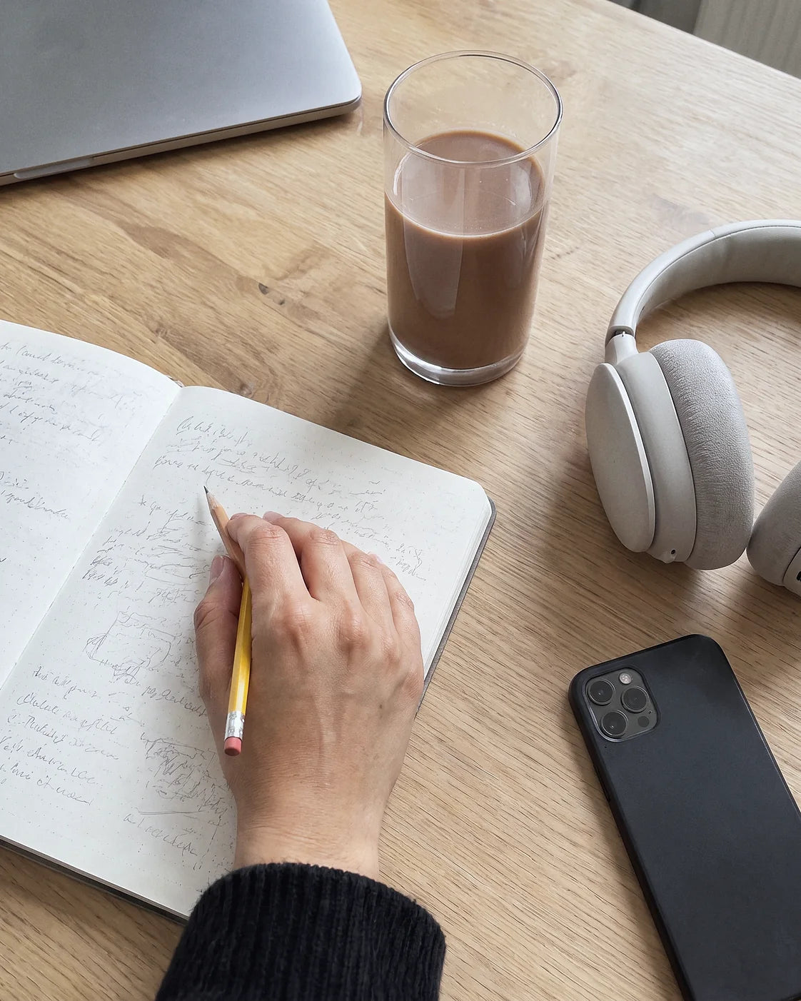 A desk seen from above: a hand writing in a notebook with a pencil, a glass of Thunder Honey, headphones and a phone