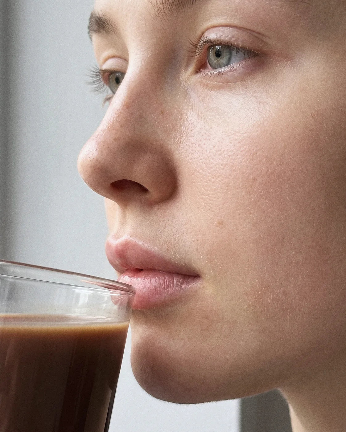 A close-up of a young woman's face as she sips Thunder Honey from a glass: clear natural skin with a few freckles