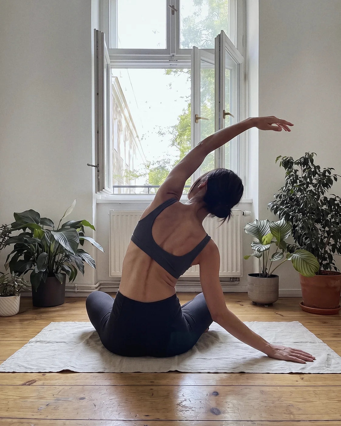 A woman seen from behind in a slow side stretch on a linen mat by an open window, plants on either side