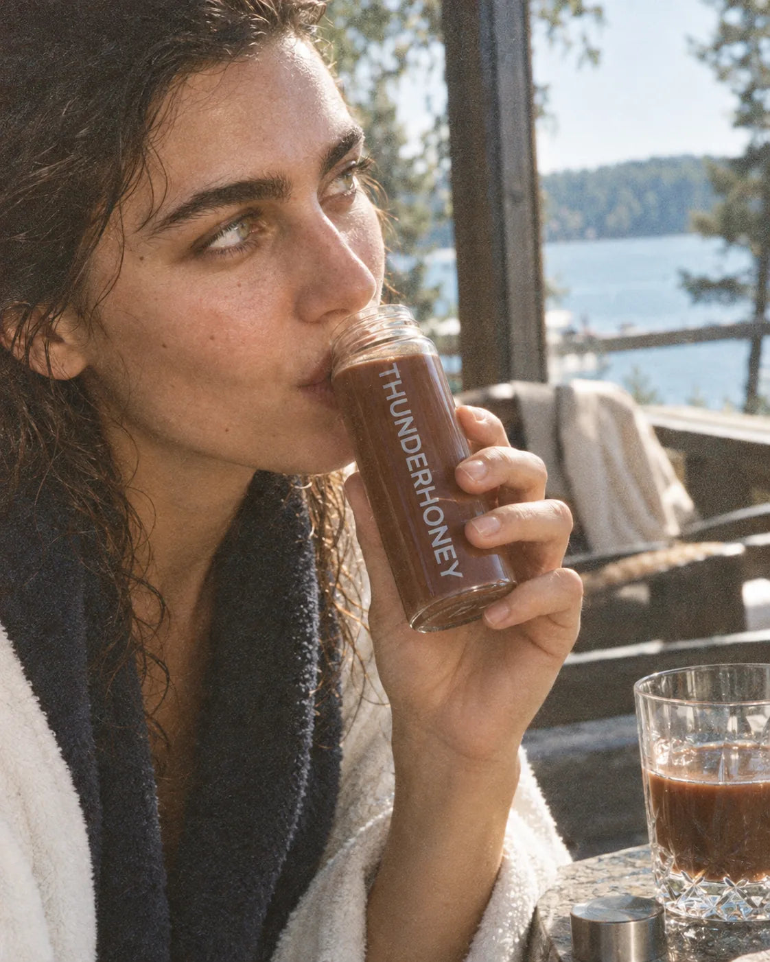 A woman in a bathrobe on a lakeside terrace, drinking Thunder Honey from the glass bottle