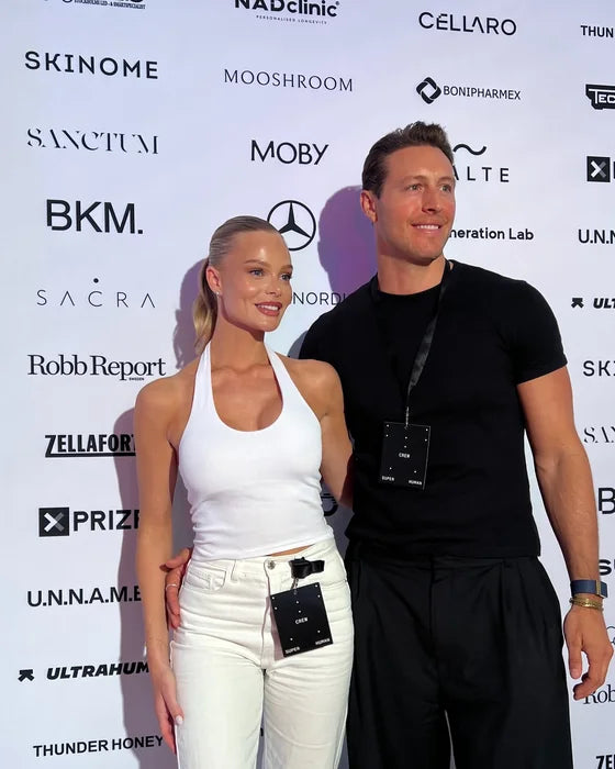 A woman in white and a man in black posing in front of the partner wall at the Superhuman event