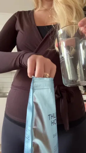 A woman in a brown wrap top holding a pouch of Thunder Honey beside an empty glass in her kitchen