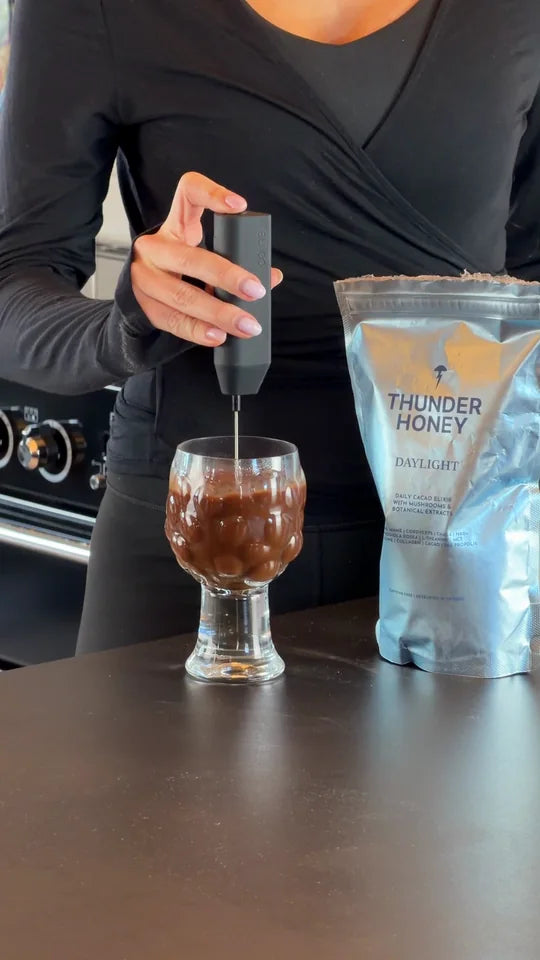 A hand frothing a glass of hot Thunder Honey with a milk frother, the silver Daylight pouch standing beside it on the counter