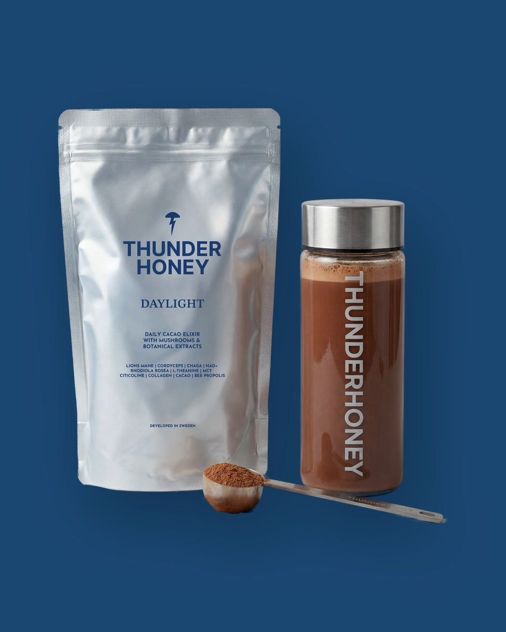 The welcome kit on deep navy: the Thunder Honey Daylight pouch, a glass bottle of made cacao with a steel cap, and a steel scoop of cacao powder